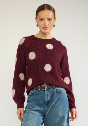 Femme portant un pull bordeaux à gros pois blancs, un jean bleu, une ceinture marron et des boucles d'oreilles créoles dorées, debout devant un fond uni.