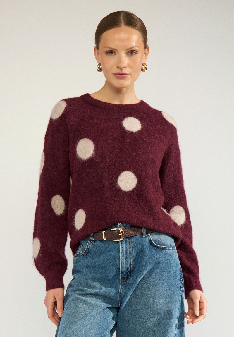 Femme portant un pull bordeaux à gros pois blancs, un jean bleu, une ceinture marron et des boucles d'oreilles créoles dorées, debout devant un fond uni.