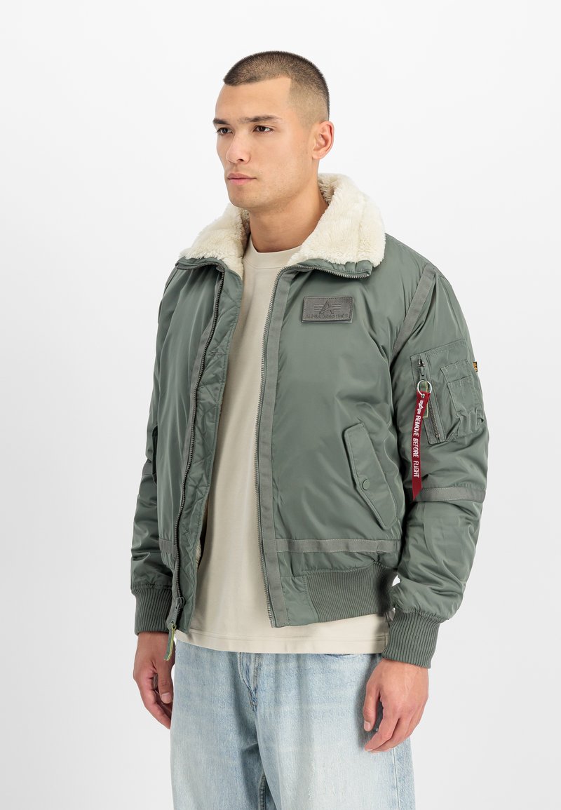 Uomo che indossa una giacca bomber verde con colletto in shearling bianco, camicia beige e jeans azzurri, in piedi su sfondo bianco.