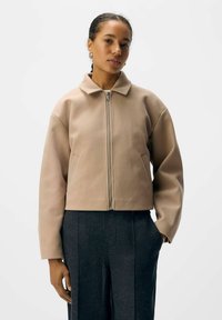 Beige lynlåsjakke med struktureret krave, drop skuldre og sidelommer, kombineret med mørke teksturerede bukser. Glat, blødt materiale.
