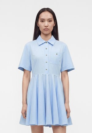 Kvinde med langt mørkt hår iført en lyseblå kortærmet kjole med krave, foran knapper, brystlomme og plisseret nederdel.