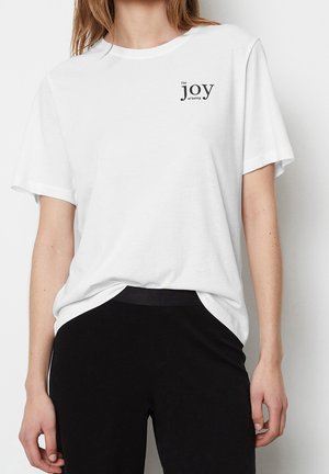 Kobieta w białej koszulce z krótkim rękawem i czarnych spodniach, z napisem „The joy of being” na piersi.