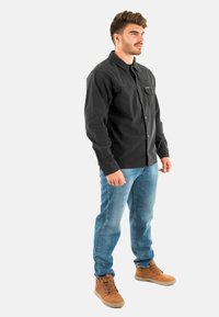 Zwarte corduroy button-up shirt met een zak, gecombineerd met blauwe denim jeans en tan laarzen, staand tegen een effen witte achtergrond.