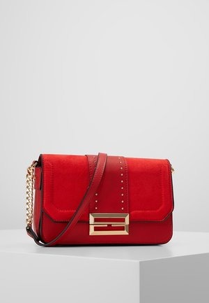 Czerwona skórzana i zamszowa torebka na ramię z złotym łańcuszkiem, dekoracyjnymi złotymi ćwiekami oraz prostokątną złotą klamrą na białej powierzchni