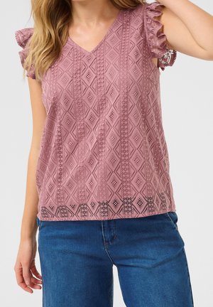 Kobieta w mauve koronkowej, bezrękawnikowej bluzce z falbankami na ramionach i niebieskich dżinsach na jednolitym tle.