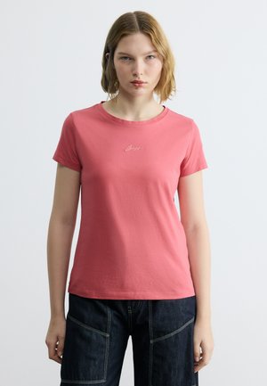 Νεαρή γυναίκα με ώμου μαλλιά ξανθά, φορώντας κοραλί ροζ κοντομάνικο πουκάμισο και σκούρο μπλε τζιν, μπροστά σε απλό φόντο.