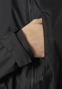 Μαύρο μπουφάν με λεία υφή, που διαθέτει τσέπη με φερμουάρ στο μανίκι και ορατό χέρι μέσα, παρουσιάζοντας λεπτομέρειες υφάσματος και σχεδίου.