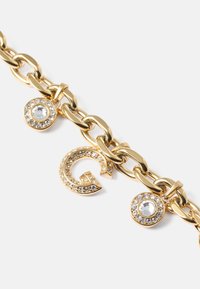 Pulseira de corrente dourada com elos grandes, apresenta uma letra 'G' cravejada de cristais e um charm circular com strass transparentes.