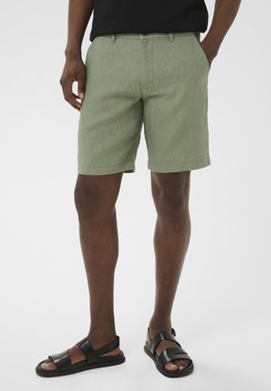 Mężczyzna w jasnooliwkowych szortach, czarnych sandałach i czarnej koszulce, stojący z rękami w kieszeniach na jednolitym tle.