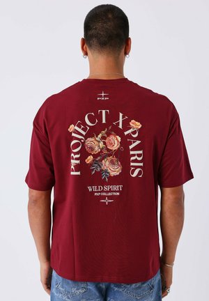Man in een bordeauxrode T-shirt met bloemontwerp en de tekst "PROJECT X PARIS WILD SPIRIT PXP COLLECTION" op de achterkant.