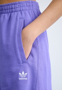 Lila bomullsbyxor med sidoficka och en vit Adidas-logotyp. Resårmidja för komfort, slät textur och avslappnad passform.