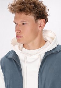 Grau melierte Fleeceweste über einem weißen Hoodie. Die Weste hat einen Reißverschluss vorne und einen hohen Kragen, der eine glatte Textur und ein weiches Erscheinungsbild zeigt.