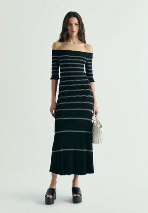 Kobieta w czarnej sukience z odkrytymi ramionami z białymi zygzakowymi paskami, trzymająca białą torebkę i nosząca czarne sandały na platformie.