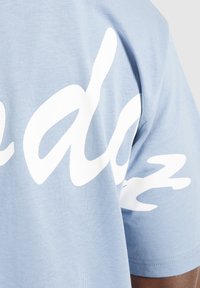 Tela azul claro con grandes letras en cursiva blancas parcialmente visibles en la manga y el área del hombro de una camisa de manga corta.
