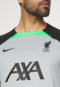Homme portant un maillot gris du Liverpool FC avec logo Nike noir, écusson noir L.F.C., grand logo de sponsor AXA noir et détail vert au col.