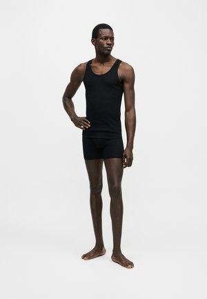 Hombre con camiseta sin mangas negra y pantalones cortos, de pie descalzo con una mano en la cadera, sobre un fondo blanco liso.