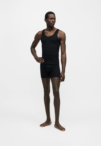 Homme en débardeur noir et short, debout pieds nus, une main sur la hanche, devant un fond blanc uni.