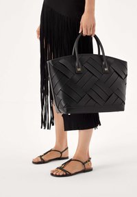 Musta kudottu nahkainen klemmarikassi, jossa on kaksi kahvaa. Siinä on jämäkkä muoto ja hillityt kultaiset yksityiskohdat. Henkilö pitää kassia mustan hapsuhameen kanssa.