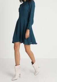 Tummansininen pitkähihaisinen mekko, jossa on levenevä helma ja nappidetalji kauluksessa, yhdistettynä valkoisiin löysiin nilkkabootseihin, joissa on terävä kärki.