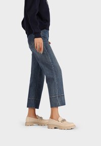 Scarpe slip-on beige con suola spessa, decorate con accessori ornamentali. Il modello indossa jeans blu scuro a gamba dritta.