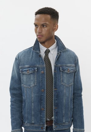 Jeansjacke in mittelblau mit zwei Brusttaschen und braunen Knöpfen, getragen über einem gestreiften Hemd und einer grün gemusterten Krawatte.