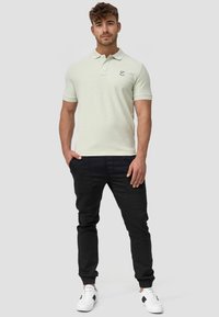 Hellgrünes Poloshirt mit Kragen und kurzen Ärmeln, mit einem Logo auf der Brust. Kombiniert mit schwarzen Jogginghosen und weißen Sneakers.