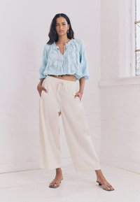 Top corto in pizzo floreale azzurro chiaro con scollatura regolabile a cordoncino, abbinato a pantaloni a gamba larga color crema e sandali neri con tacco.