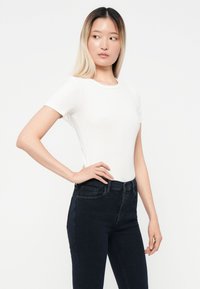 Fehér, rövid ujjú, testhez álló póló bordás textúrával, sötétkék, magas derekú farmernadrággal párosítva, amely egy laza megjelenést mutat.