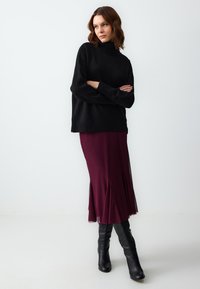 Pull en maille noire à col roulé, associé à une jupe fluide bordeaux foncé dotée de fentes latérales, et des bottes en cuir noires jusqu'aux genoux.