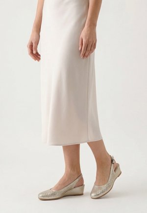 Kvinde iført lys beige midi nederdel og beige lave kilehælssandaler med perforeret design, der står på en hvid gulv.