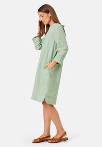 Jasnozielona lniana sukienka z rękawami o długości trzech czwartych, bocznymi kieszeniami oraz falbaniastym dołem; połączona z brązowymi sandałami na paski.
