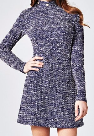 Kobieta ubrana w dopasowaną, długorękawową sukienkę z wysokim kołnierzem, wykonaną z niebiesko-białej dzianiny o teksturowanym wzorze, z perłowymi guzikami na kołnierzu.