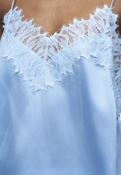 Ανοιχτό γαλάζιο σατέν καμιζόλ με λεπτή λευκή δαντέλα κατά μήκος του V-λαιμού και λεπτές ιμάντες στους ώμους πάνω σε μαυρισμένο δέρμα.