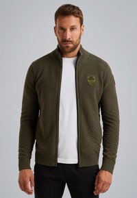 Giacca zip con maniche lunghe di colore verde oliva, caratterizzata da un motivo a maglia testurizzata, polsini e orlo a coste, e una toppa con logo sul petto. Indossata sopra una camicia bianca.
