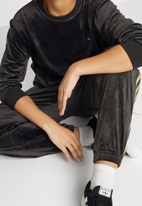 Osoba v černém sametovém teplákovém kostýmu se žlutými pruhy, v bílých ponožkách a černých teniskách Adidas, sedící na podlaze s jedním kolenem vztyčeným.