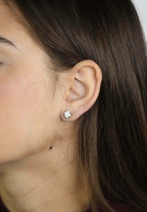 Vrouw met lang bruin haar die een enkele briljant geslepen diamant oorbel draagt in haar linkeroor tegen een neutrale achtergrond.