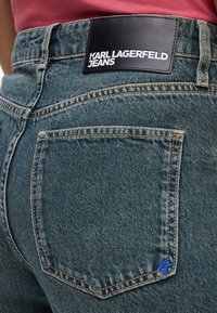 Sötétkék mosású farmernadrág, fekete bőr címkével, amelyen a "KARL LAGERFELD JEANS" felirat található, és kék hímzett részlet a zseben.
