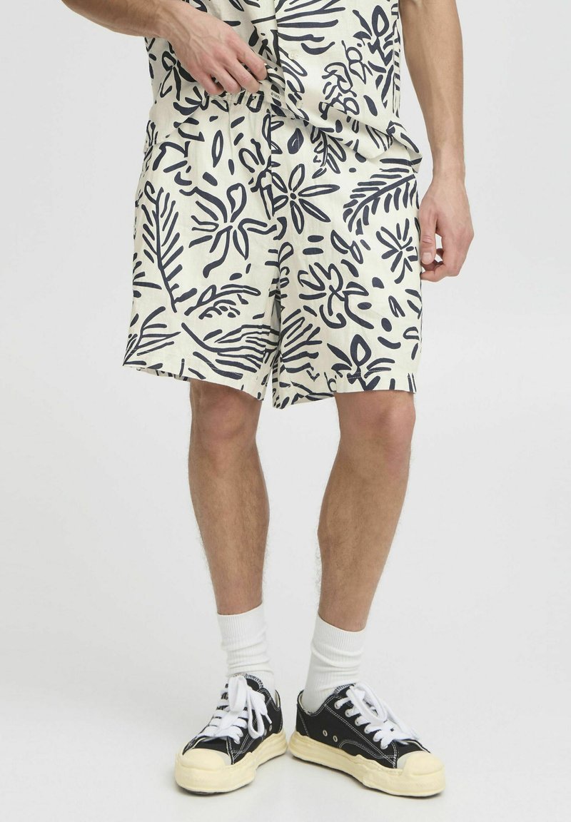 Pantalones cortos crema con estampado botánico en azul marino, cinturilla elástica, bolsillos laterales y un corte holgado, combinados con zapatillas negras y calcetines blancos.