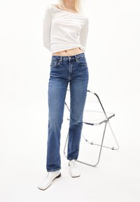 Vaqueros azules de mezclilla de talle alto con corte recto, cinco bolsillos y costuras claras, combinados con una camisa blanca de manga larga y zapatos blancos.