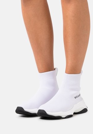Jambes portant des baskets style chaussettes blanches avec des semelles noires et le texte « BULLBOXER » sur le côté, sur un fond neutre.