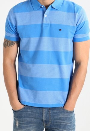 Mann trägt blau gestreiftes Poloshirt und blaue Jeans mit Händen in den Taschen, Tätowierung sichtbar am linken Arm.