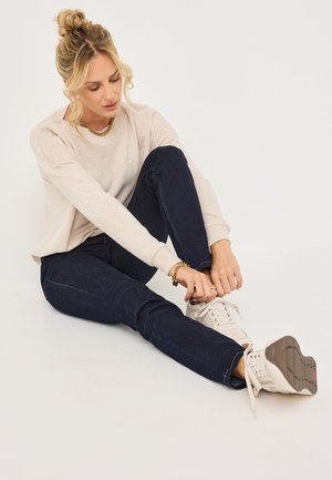 Frau mit blonden Haaren im Dutt, die einen beigen Pullover, dunkle Jeans und weiße Sneakers trägt und auf dem Boden sitzt, um ihren Schuh zu binden.