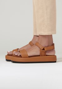 Brown leather sandals with wide straps, cushioned insoles, and a thick platform sole. The design includes adjustable ankle straps.