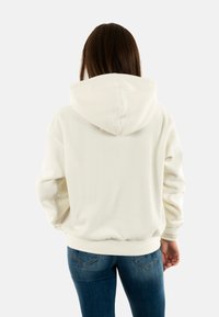 Sweat-shirt à capuche blanc avec texture côtelée, coupe décontractée et poignets. La vue arrière montre un design simple et un tissu doux. Associé à un jean bleu.