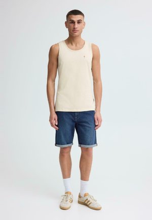 Mężczyzna stojący w beżowej koszulce na ramiączkach, podwiniętych dżinsowych szortach, białych skarpetkach i beżowych trampkach na tle jasnego, jednolitego tła.