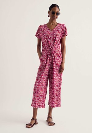 Woman wearing pink floral wide-leg jumpsuit with short sleeves, tie waist, sunglasses, cherry earrings, and brown sandals, standing relaxed.