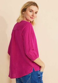 Blusa fucsia, comoda con maniche a tre quarti, orlo arrotondato e dettagli sottili con piega sul retro. Indossata con jeans blu.