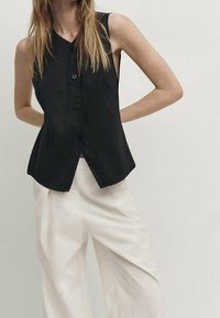 Gilet nero senza maniche con scollatura a V, dotato di chiusura con bottone e silhouette affusolata, abbinato a pantaloni bianchi morbidi. Tessuto dalla consistenza liscia.