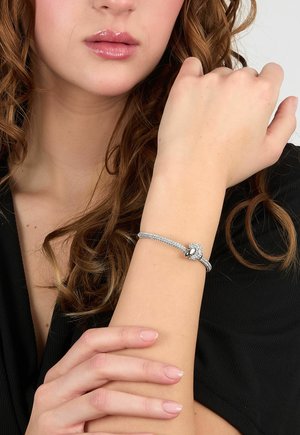 Donna con capelli ricci che indossa un top nero mostra un braccialetto d'argento con dettagli scintillanti sul polso sinistro sollevato.