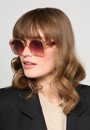 Donna con capelli castano chiaro mossi, che indossa grandi occhiali da sole rosa, blazer nero e top beige, guarda leggermente di lato.